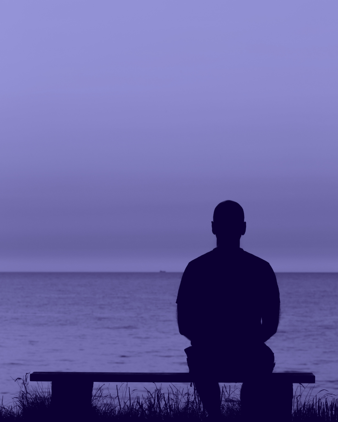 Silhouette of a person sitting on a bench by the water, facing the horizon at dusk.